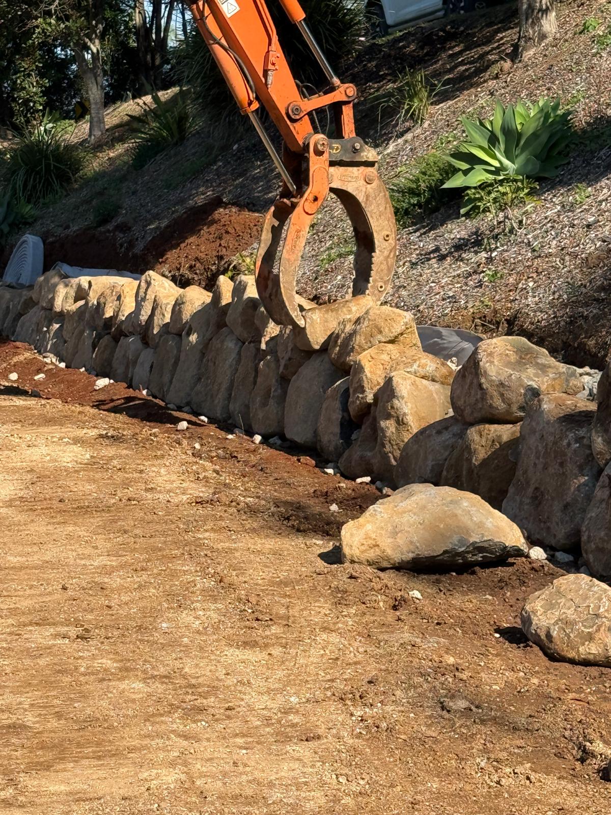 Excavator grab placing graded rock along a retaining wall on site
